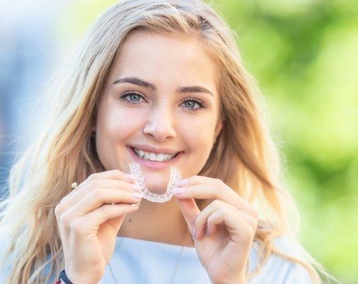 Young blonde woman smiling and holding Invisalign clear aligner outdoors