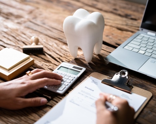 Person typing on calculator and writing on clipboard on desk with model of tooth