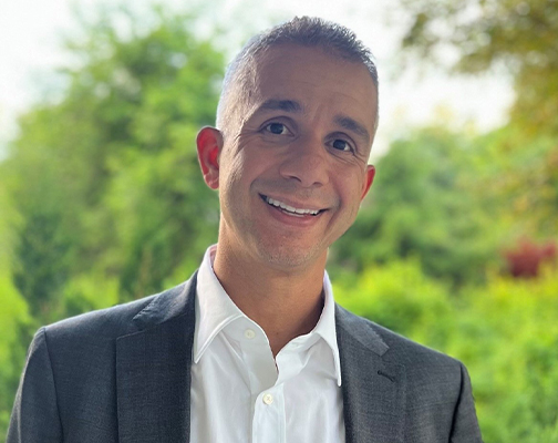 Joe wearing gray suit jacket and white dress shirt outdoors with green trees in background