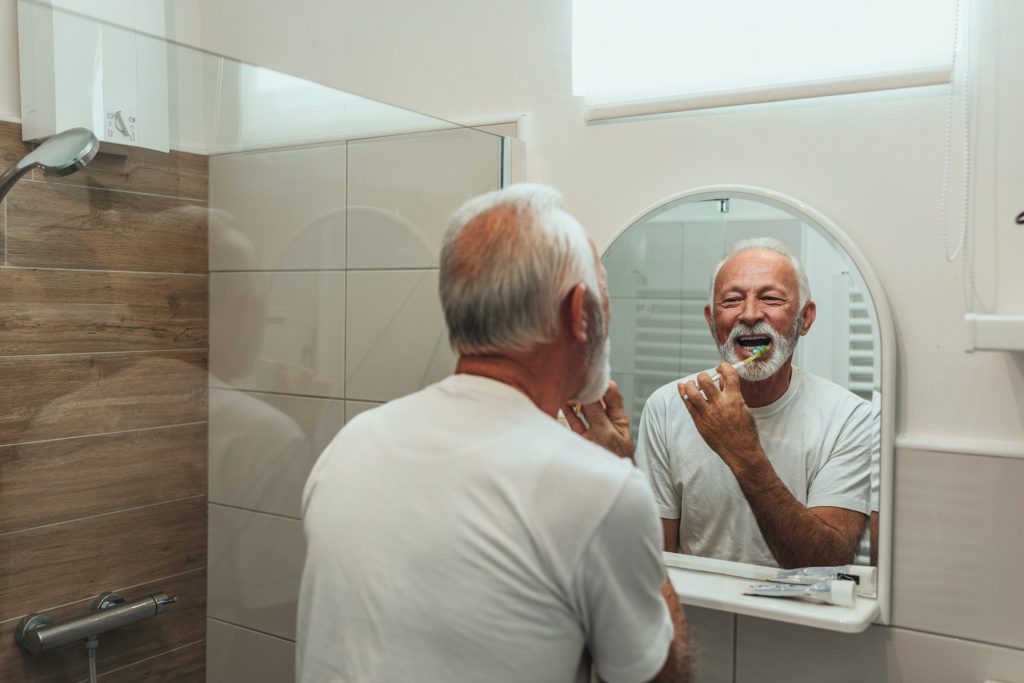 Senior man smiling in bathroom mirror while brushing his teeth