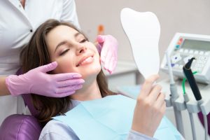 Woman with brown hair in purple dentists chair holding a tooth shaped mirror while the dentist stands behind her